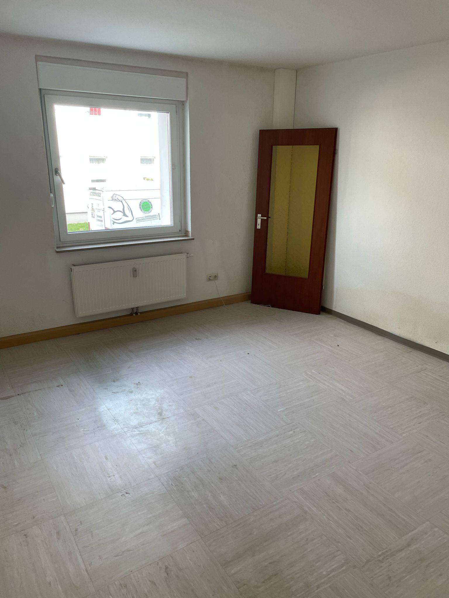 Nach einer erfolgreichen Entrümpelung präsentiert sich dieser Raum in neuem Glanz: Weiße Wände schaffen ein freundliches Ambiente, während ein großes Fenster mit einem darunterliegenden Heizkörper für ausreichend Tageslicht sorgt. Wo einst noch Möbel und Gegenstände standen, herrscht jetzt Ordnung - lediglich eine Holztür mit gelb getöntem Glas lehnt an der Wand und wartet auf den nächsten Einsatz. Der Boden ist dank heller Fliesen leicht zu reinigen und wirkt modern sowie gepflegt. So bietet Ihnen die gründliche Entrümpelung von Pottentruempler.de die perfekte Grundlage für eine neue Nutzung Ihrer Räume. ---.