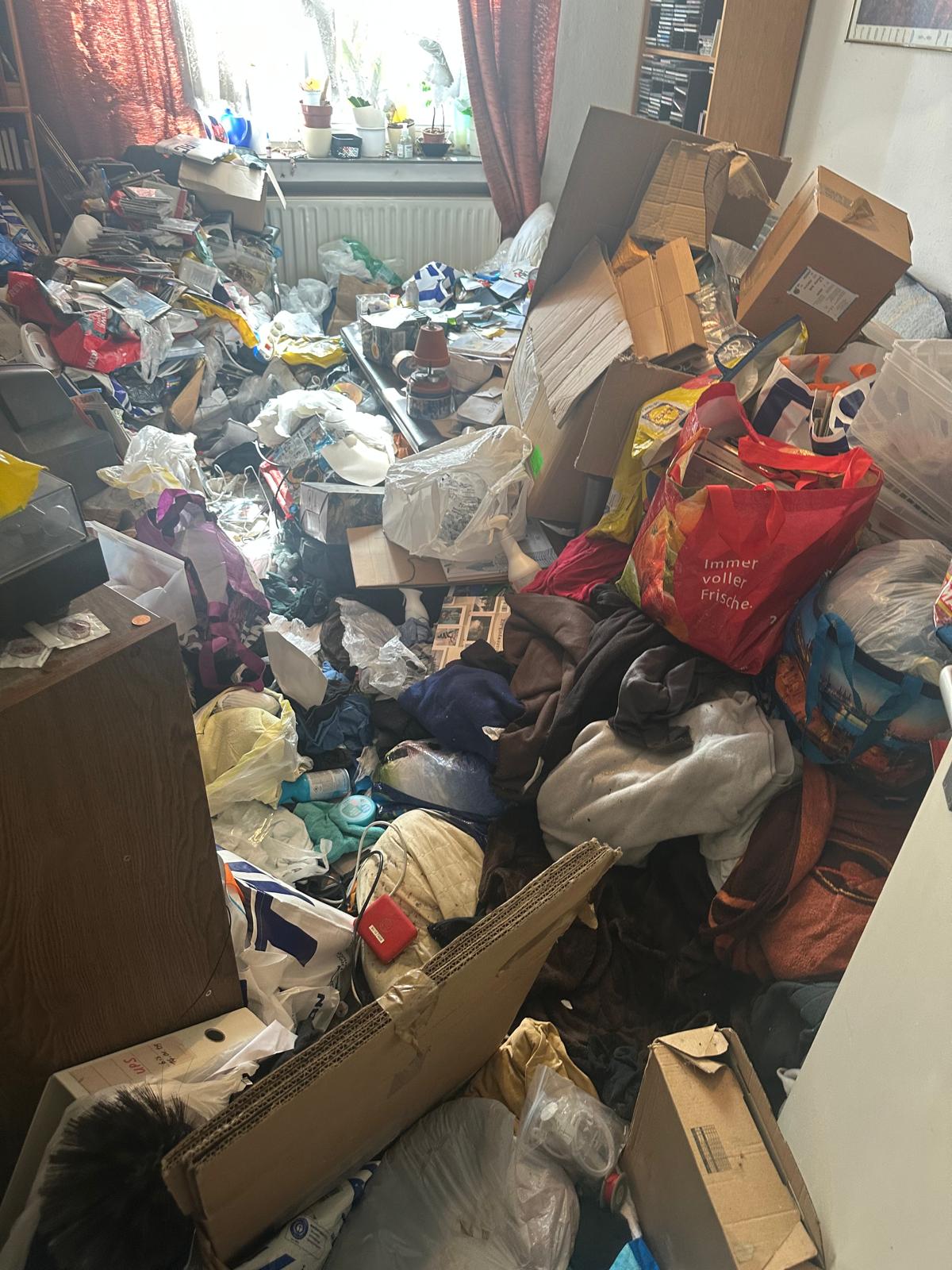 Ein unordentlicher Raum mit Stapeln von Kleidern, Taschen, Kisten und anderen Gegenständen, die den Boden bedecken, während das Sonnenlicht durch ein Fenster fällt, das teilweise durch rote Vorhänge verdeckt ist. Ein unordentlicher Raum mit Stapeln von Kleidern, Taschen, Kisten und anderen Gegenständen, die den Boden bedecken, während das Sonnenlicht durch ein Fenster fällt, das teilweise durch rote Vorhänge verdeckt ist.