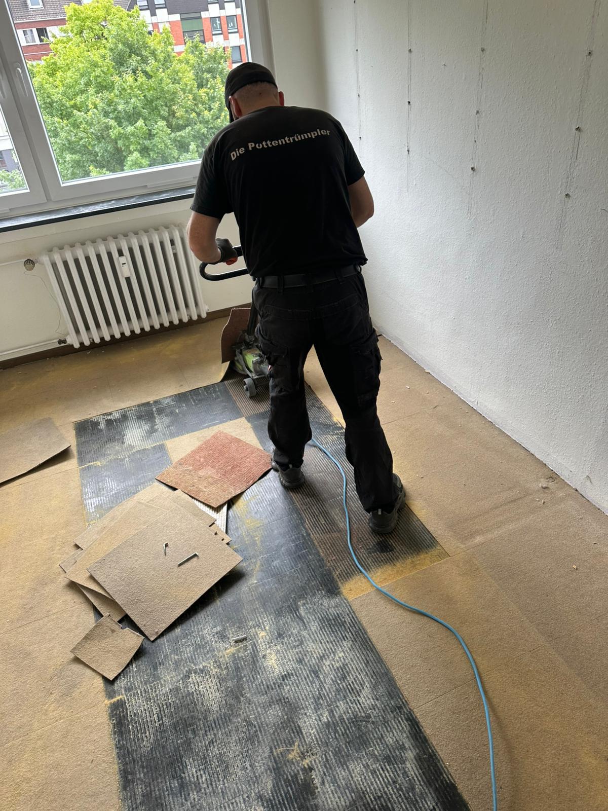 Eine Person in Arbeitskleidung entfernt mit einem Elektrowerkzeug den alten Bodenbelag in einem Raum. Teile des Bodenbelags liegen verstreut auf dem Boden, und im Hintergrund ist ein großes Fenster mit Blick auf Bäume zu sehen. Eine Person in Arbeitskleidung entfernt mit einem Elektrowerkzeug den alten Bodenbelag in einem Raum. Teile des Bodenbelags liegen verstreut auf dem Boden, und im Hintergrund ist ein großes Fenster mit Blick auf Bäume zu sehen.