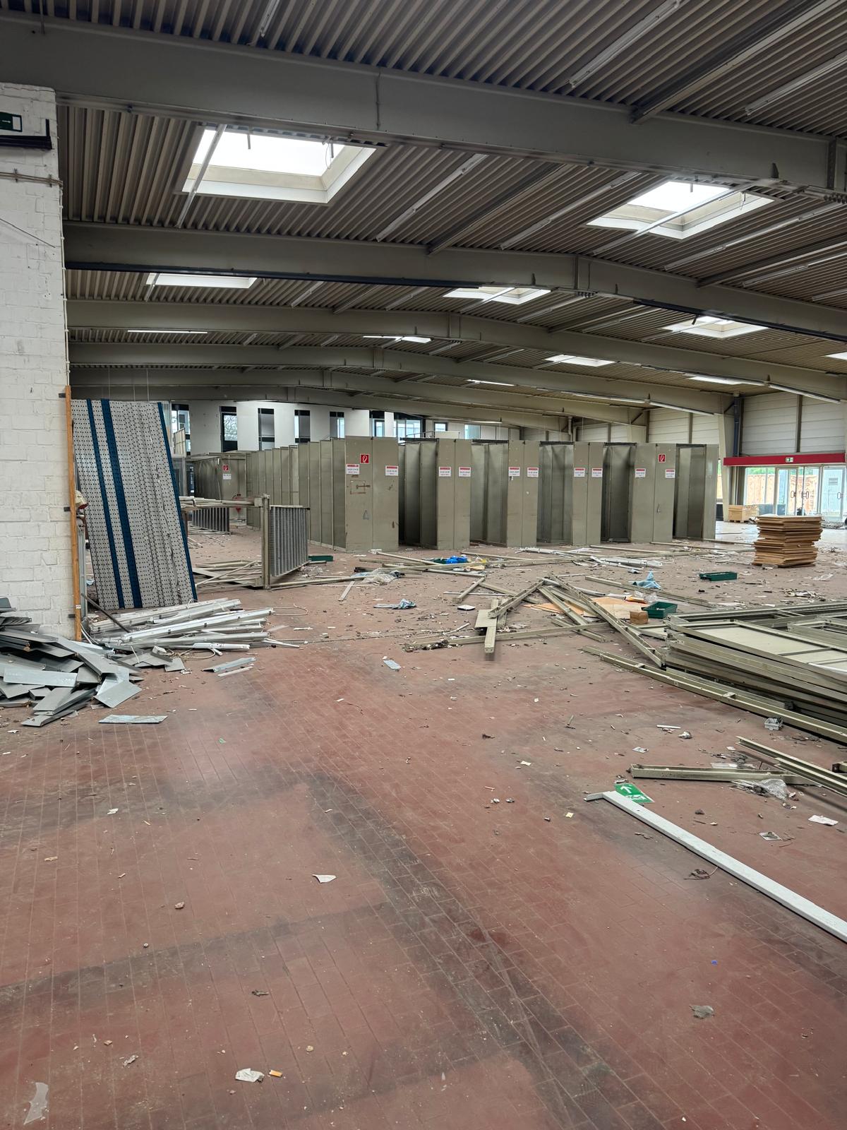 Ein leeres, großes Industrielager mit einem unordentlichen, mit Schutt und Baumaterialien bedeckten Boden. Metallschränke säumen die Rückseite, während Oberlichter an der Decke natürliches Licht hereinlassen. Ein leeres, großes Industrielager mit einem unordentlichen, mit Schutt und Baumaterialien bedeckten Boden. Metallschränke säumen die Rückseite, während Oberlichter an der Decke natürliches Licht hereinlassen.
