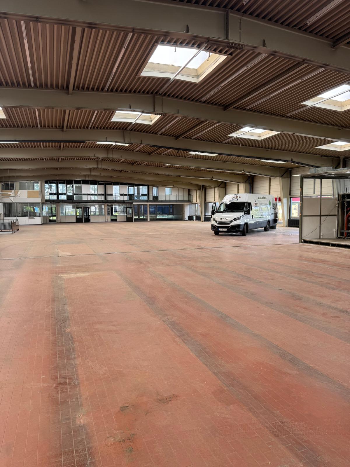 Ein großes, größtenteils leeres Lagerhaus mit einem rot gefliesten Boden, hohen Decken, Oberlichtern und einem weißen Lieferwagen, der in der Mitte geparkt ist. Natürliches Licht fällt durch das Dach und die Glasfenster auf der Rückseite ein. Ein großes, größtenteils leeres Lagerhaus mit einem rot gefliesten Boden, hohen Decken, Oberlichtern und einem weißen Lieferwagen, der in der Mitte geparkt ist. Natürliches Licht fällt durch das Dach und die Glasfenster auf der Rückseite ein.